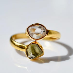 Peut inclure: Une bague ouverte en or ornée de deux pierres distinctes : une pierre transparente à facettes de forme ovale et une pierre brute de couleur vert olive aux formes irrégulières. L'anneau est en or lisse et poli, créant un design ouvert.
