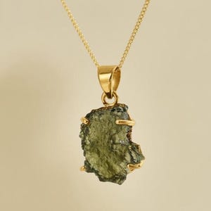 May include: A gold-colored necklace featuring a raw, olive-green gemstone pendant. The pendant is irregularly shaped with a textured surface and is held in place by a gold-colored metal setting. The chain is also gold-colored.