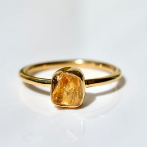 May include: A gold ring featuring a single, raw, amber-colored gemstone set in a polished metal band. The stone has an irregular, faceted shape with a translucent, warm yellow-orange hue, contrasting with the smooth, reflective gold-toned band.