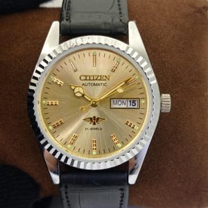 May include: A Citizen automatic wristwatch with a gold-toned face, featuring the day and date. The watch has a silver-colored bezel and a black leather band. The watch face displays the text "CITIZEN AUTOMATIC" and "MON 15".