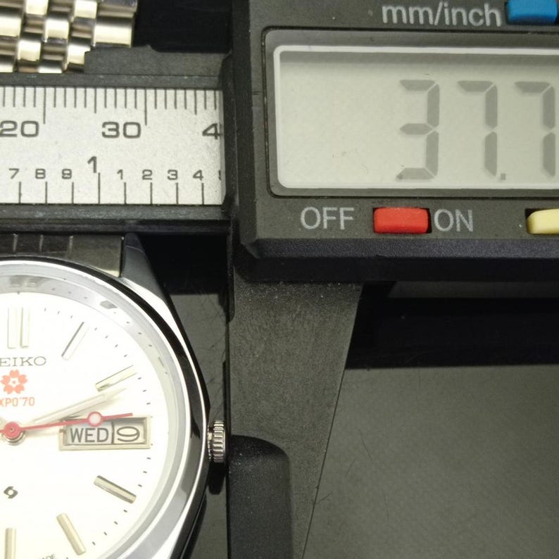 May include: A silver-tone Seiko wristwatch with a white dial, featuring a red second hand and a day-date window displaying WED 9. A digital caliper shows a measurement of 37.7 mm. The watch has a metallic link bracelet and a cherry blossom logo.