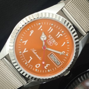 Könnte beinhalten: Eine Seiko Automatik-Armbanduhr mit orangefarbenem Zifferblatt und arabischen Ziffern. Die Uhr hat ein silberfarbenes, strukturiertes Armband und eine silberne Lünette. Tag und Datum werden in einem weißen Rechteck angezeigt. Die Zeiger sind silber und rot.