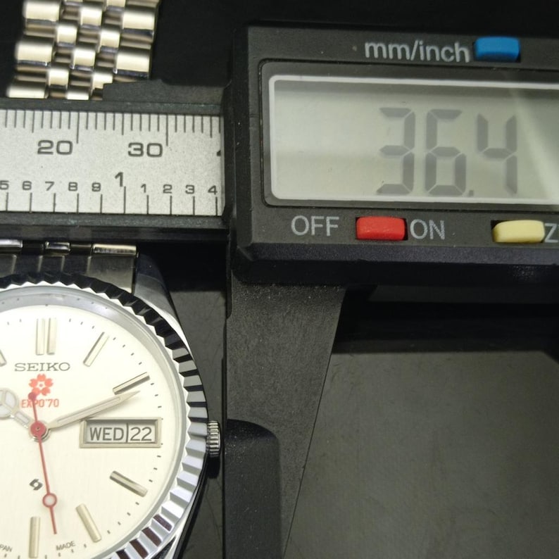 May include: A silver-tone Seiko wristwatch with a white dial, featuring a red flower logo and the text EXPO '70. The watch has a fluted bezel, a day-date window, and a metal link bracelet. A digital caliper shows a measurement of 36.4 mm.