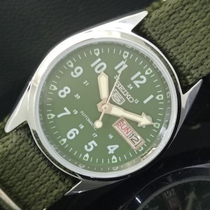 Könnte beinhalten: Nahaufnahme einer Seiko Automatik-Armbanduhr mit grünem Zifferblatt und silberner Lünette. Die Uhr hat weiße Ziffern und Zeiger, mit Tages- und Datumsanzeige. Das Armband ist aus grünem Stoff.