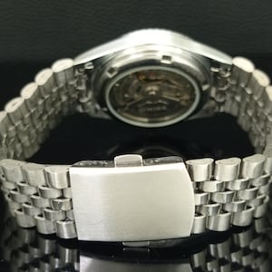K&ouml;nnte beinhalten: Eine silberfarbene Armbanduhr aus Edelstahl mit einem mehrgliedrigen Armband und einer Faltschlie&szlig;e. Der Geh&auml;useboden der Uhr ist transparent und gibt den Blick auf das mechanische Uhrwerk und die Zahnr&auml;der in kreisf&ouml;rmiger Anordnung frei.