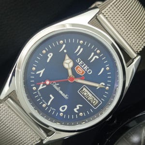 May include: A stainless steel Seiko 5 automatic watch with a blue dial and Arabic numerals. The watch has a silver mesh strap and a day-date display. The dial reads "Automatic" and "Made in Japan".