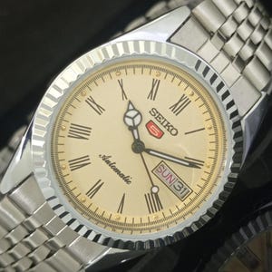 May include: A silver Seiko automatic wristwatch with a champagne-colored face. The watch features Roman numeral hour markers, a day-date display, and a silver band. The watch face has the word "Automatic" printed on it.