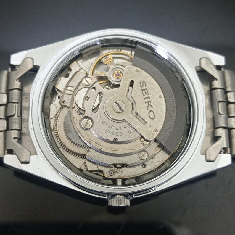 K&ouml;nnte beinhalten: Eine silberfarbene Armbanduhr mit abgenommenem Geh&auml;useboden, die das interne mechanische Uhrwerk freilegt. Die Schwungmasse ist mit dem Text SEIKO, 6309A und 17 JEWELS graviert. Die Uhr verf&uuml;gt &uuml;ber ein metallisches Gliederarmband.
