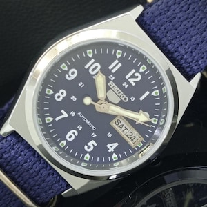 Könnte beinhalten: Eine silberfarbene Seiko Automatik-Armbanduhr mit marineblauem Zifferblatt und blauem Stoffarmband. Das Zifferblatt zeigt weiße Ziffern, Zeiger und den Wochentag. Das Datum ist Samstag, der 24.