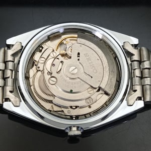 May include: A silver-toned wristwatch with the back casing removed to reveal the internal mechanical movement. The metal oscillating weight is engraved with the text SEIKO and 6309A 17 JEWELS. A metallic link bracelet is attached to the watch case.