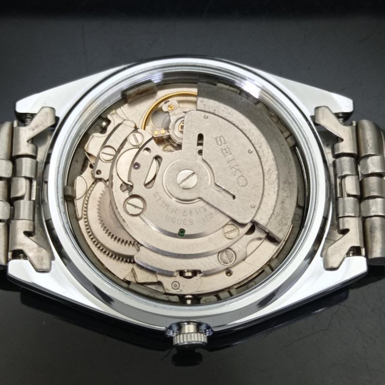 May include: A silver-toned wristwatch with the back casing removed to reveal the internal mechanical movement. The metal oscillating weight is engraved with the text SEIKO and 6309A 17 JEWELS. A metallic link bracelet is attached to the watch case.