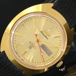 Puede incluir: Un reloj de pulsera dorado con una esfera texturizada, con el texto "HMT Konchon" y "AUTOMATIC 21 JEWELS". El reloj tiene una pantalla de día y fecha y está colocado sobre una correa negra. El reloj está marcado "MADE IN 021A INDIA".