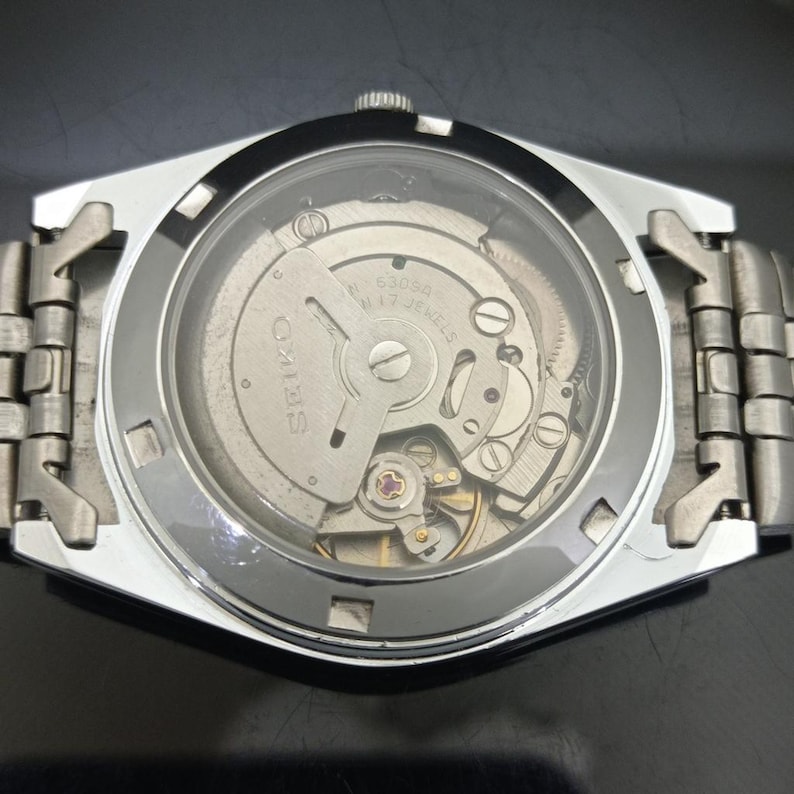May include: A silver-toned wristwatch with an exhibition case back revealing the internal mechanical movement. The rotor is engraved with the text SEIKO, 6309A, and 17 JEWELS. The watch features a metallic link bracelet and a circular glass window.