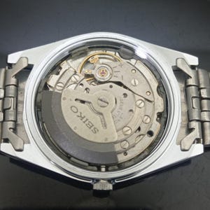 K&ouml;nnte beinhalten: Ein silberfarbenes mechanisches Uhrwerk mit abgenommenem Geh&auml;useboden, das die internen Zahnr&auml;der und eine graue Schwungmasse freilegt. Der Metallrotor ist mit dem Wort SEIKO und dem Text SEVENTEEN JEWELS JAPAN graviert.