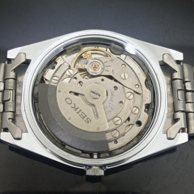 K&ouml;nnte beinhalten: Ein silberfarbenes mechanisches Uhrwerk mit abgenommenem Geh&auml;useboden, das die internen Zahnr&auml;der und eine graue Schwungmasse freilegt. Der Metallrotor ist mit dem Wort SEIKO und dem Text SEVENTEEN JEWELS JAPAN graviert.