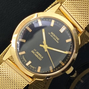 May include: A gold-toned wristwatch with a black face. The watch features gold hands and markers, and a mesh band. The watch face reads "MONTINE OF SWITZERLAND 17 JEWELS INCABLOC SWISS MADE".