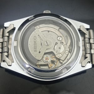 K&ouml;nnte beinhalten: Eine silberfarbene Seiko-Armbanduhr mit einem Sichtboden, der das interne mechanische Uhrwerk freilegt. Der Rotor ist mit dem Text Seiko 6309A 17 Jewels graviert. Die Uhr verf&uuml;gt &uuml;ber ein Metallgliederarmband und ein kreisf&ouml;rmiges Glasfenster.