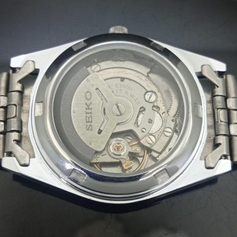 K&ouml;nnte beinhalten: Eine silberfarbene Seiko-Armbanduhr mit einem Sichtboden, der das interne mechanische Uhrwerk freilegt. Der Rotor ist mit dem Text Seiko 6309A 17 Jewels graviert. Die Uhr verf&uuml;gt &uuml;ber ein Metallgliederarmband und ein kreisf&ouml;rmiges Glasfenster.