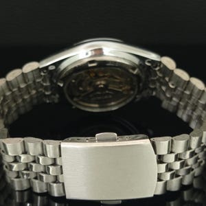 May include: A silver-toned metal wristwatch featuring a multi-link bracelet with a fold-over clasp. The watch case back is transparent, revealing the internal mechanical movement and gears, with the brand name Seiko visible on the rotor.