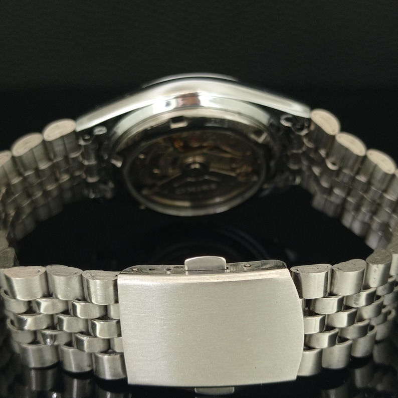 May include: A silver-toned metal wristwatch featuring a multi-link bracelet with a fold-over clasp. The watch case back is transparent, revealing the internal mechanical movement and gears, with the brand name Seiko visible on the rotor.