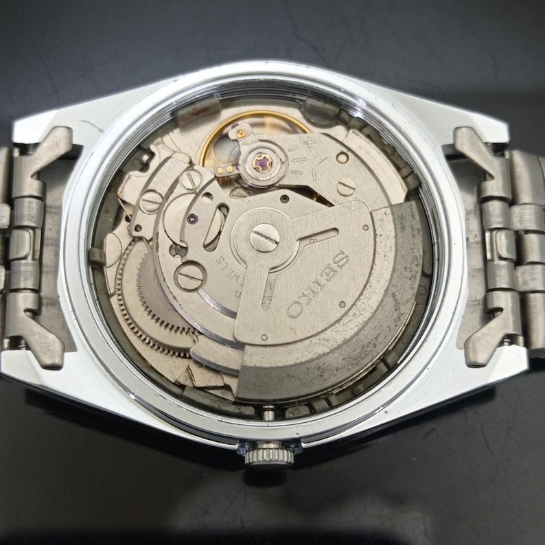 May include: A silver-toned wristwatch with an exposed mechanical movement visible through the open case back. The internal rotor is engraved with the word SEIKO and features visible gears, springs, and a balance wheel within the metallic housing.