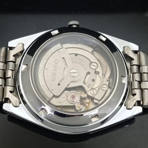 May include: A silver-toned Seiko wristwatch featuring an exhibition case back that reveals the internal mechanical movement. The watch has a metallic link bracelet and a circular glass window on the back, displaying the gears and the Seiko logo.