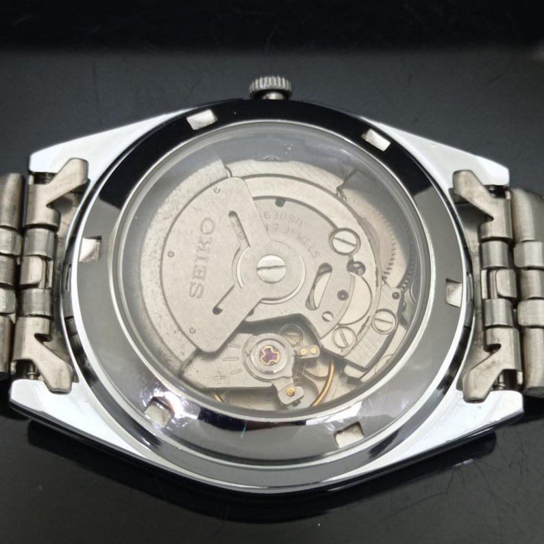 May include: A silver-toned Seiko wristwatch featuring an exhibition case back that reveals the internal mechanical movement. The watch has a metallic link bracelet and a circular glass window on the back, displaying the gears and the Seiko logo.