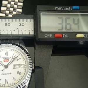 K&ouml;nnte beinhalten: Eine silberfarbene Seiko 5 Automatikuhr mit wei&szlig;em Zifferblatt, r&ouml;mischen Ziffern und einer Tages-Datums-Anzeige. Ein digitaler Messschieber zeigt ein Ma&szlig; von 36,4 mm f&uuml;r das Uhrengeh&auml;use. Die Uhr verf&uuml;gt &uuml;ber ein metallisches Gliederarmband.