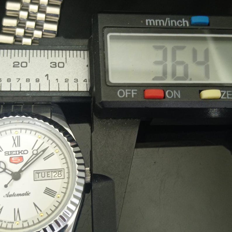 K&ouml;nnte beinhalten: Eine silberfarbene Seiko 5 Automatikuhr mit wei&szlig;em Zifferblatt, r&ouml;mischen Ziffern und einer Tages-Datums-Anzeige. Ein digitaler Messschieber zeigt ein Ma&szlig; von 36,4 mm f&uuml;r das Uhrengeh&auml;use. Die Uhr verf&uuml;gt &uuml;ber ein metallisches Gliederarmband.
