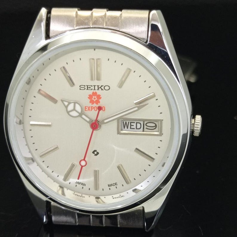 May include: A silver-tone Seiko wristwatch with a cream-colored dial, featuring a red cherry blossom logo above the text EXPO 90. The watch includes silver hour markers, a red second hand, and a day-date window displaying WED 9 at the 3 o'clock position.