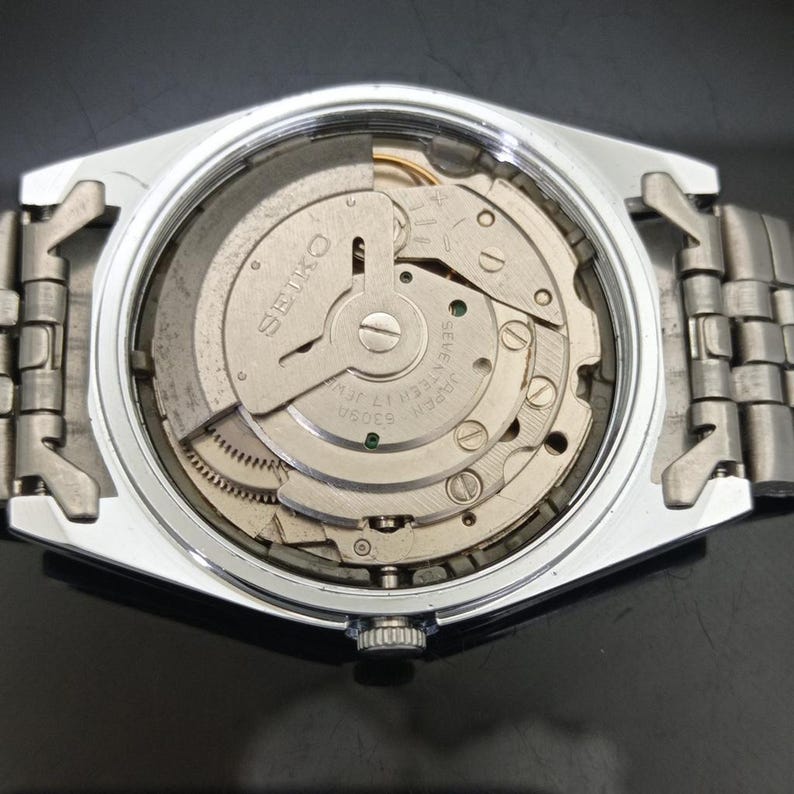 May include: A silver-toned wristwatch with the back casing removed to reveal the internal mechanical movement. The metal rotor is engraved with the text SEIKO, JAPAN, and 6309A SEVENTEEN 17 JEWELS. The watch features a metallic link bracelet.