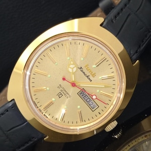Puede incluir: Un reloj de pulsera dorado con una correa de cuero negro. La esfera muestra el día y la fecha, con el texto "hmt Kanchan" y "Automatic 21 Jewels". El reloj tiene un bisel dorado y una esfera dorada.