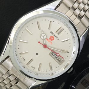 May include: A silver-tone Seiko wristwatch with a white dial featuring the text SEIKO, EXPO '70, and a red cherry blossom graphic. The watch includes a day-date window at the three o'clock position, silver hour markers, and a metal link bracelet.