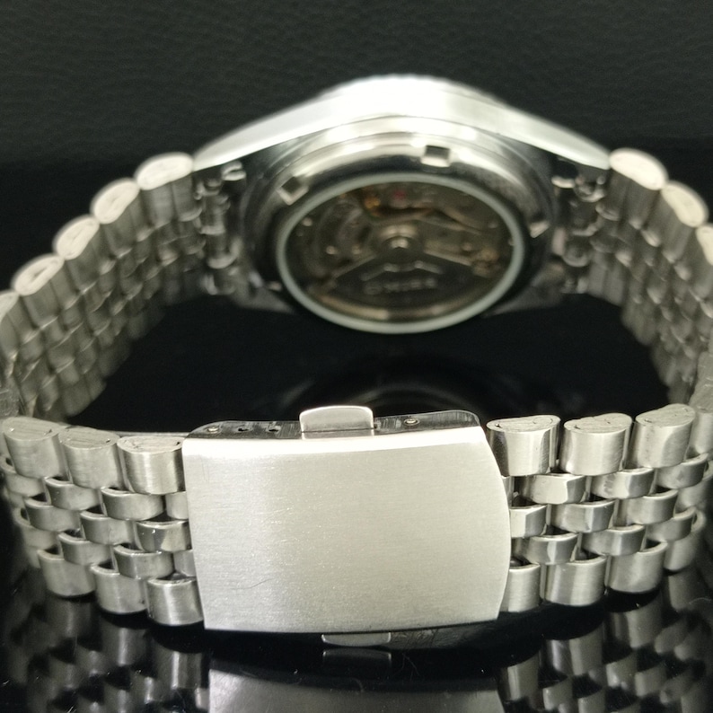 May include: A silver-toned stainless steel wristwatch featuring a multi-link bracelet with a fold-over clasp. The watch case back is transparent, revealing the internal mechanical movement and gears of the timepiece in a circular configuration.