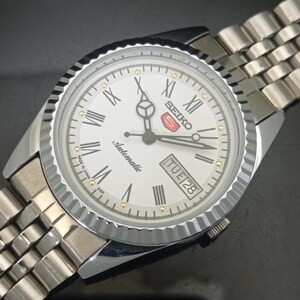 K&ouml;nnte beinhalten: Eine silberfarbene Seiko 5 Automatikuhr mit wei&szlig;em Zifferblatt, schwarzen r&ouml;mischen Ziffern als Stundenmarkierungen und einer geriffelten L&uuml;nette. Die Uhr verf&uuml;gt &uuml;ber eine Datums- und Wochentagsanzeige bei drei Uhr, Leuchtzeiger und ein Metallarmband.