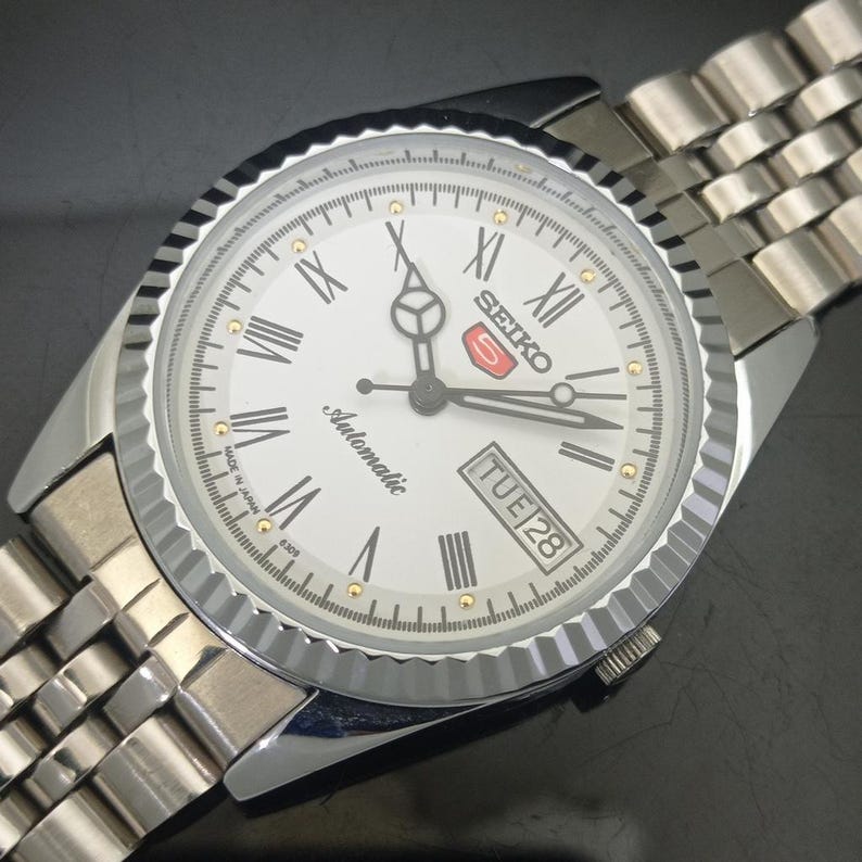 K&ouml;nnte beinhalten: Eine silberfarbene Seiko 5 Automatikuhr mit wei&szlig;em Zifferblatt, schwarzen r&ouml;mischen Ziffern als Stundenmarkierungen und einer geriffelten L&uuml;nette. Die Uhr verf&uuml;gt &uuml;ber eine Datums- und Wochentagsanzeige bei drei Uhr, Leuchtzeiger und ein Metallarmband.