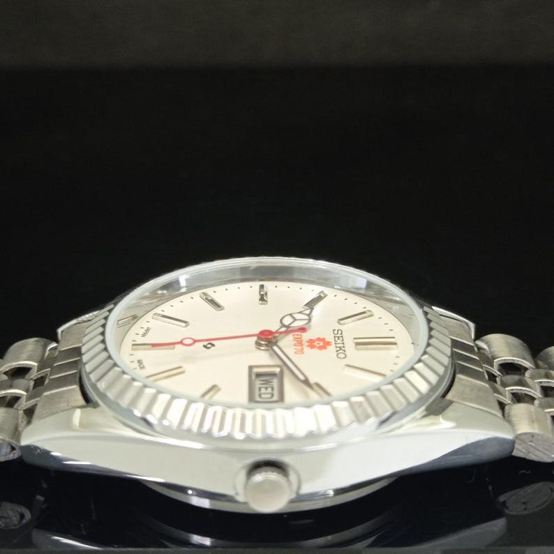 May include: A silver-tone Seiko wristwatch with a white dial, a fluted bezel, and a metal link bracelet. The dial features a red second hand, a day-date window at the three o'clock position, and the text Seiko and Expo 70 printed in red.