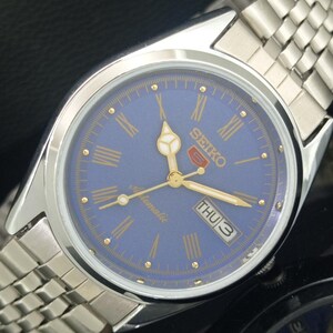 May include: A silver-toned Seiko 5 automatic watch with a blue face. The watch features gold-colored hands and hour markers, Roman numerals, and a day-date display. The watch has a silver-toned metal band.