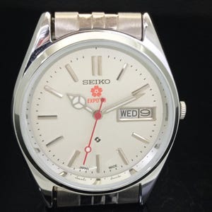 May include: A silver-tone Seiko wristwatch with a white dial, featuring a red second hand, a day-date window at the three o'clock position, and a cherry blossom graphic above the text EXPO '90. The watch includes a metallic link bracelet.