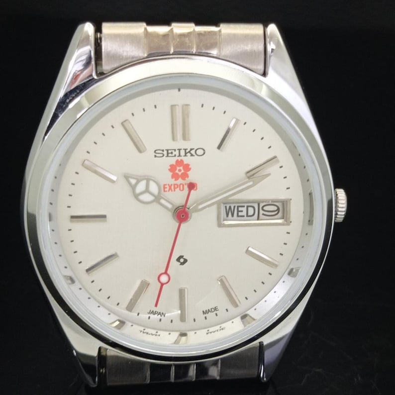 May include: A silver-tone Seiko wristwatch with a white dial, featuring a red second hand, a day-date window at the three o'clock position, and a cherry blossom graphic above the text EXPO '90. The watch includes a metallic link bracelet.