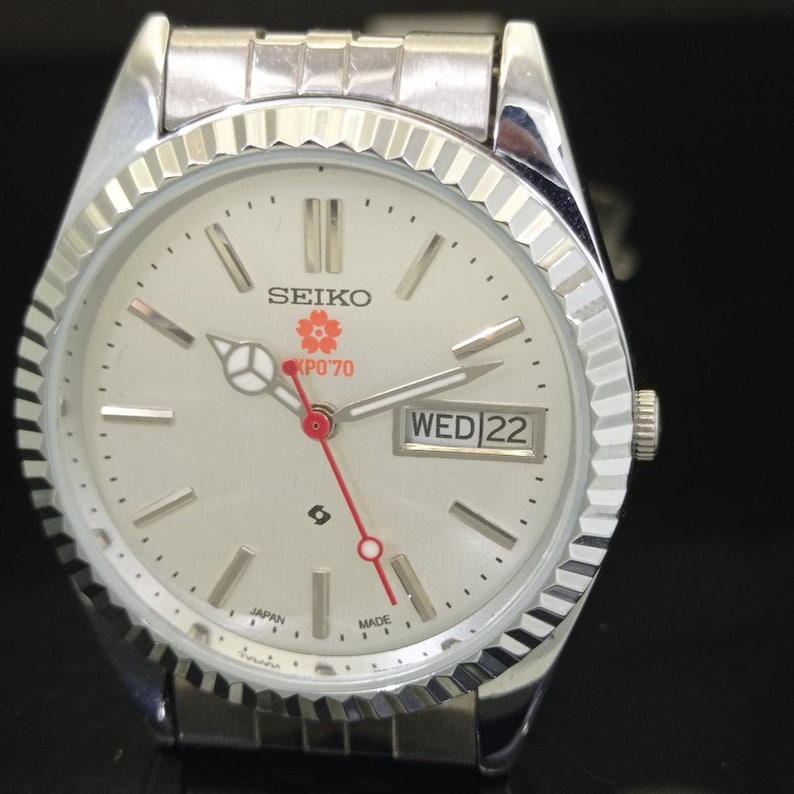 May include: A silver-tone Seiko wristwatch with a fluted bezel and a white dial. The dial features silver baton indices, a day-date window at the three o'clock position, a red second hand, and a red cherry blossom logo with the text EXPO'70.