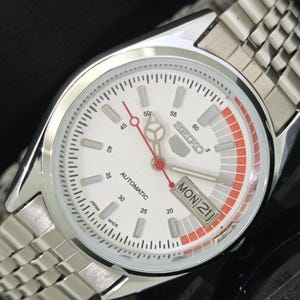 May include: A silver-tone Seiko 5 automatic wristwatch with a white dial, a red second hand, and a day-date window at the three o'clock position. The dial features a red-accented minute track, silver hour markers, and a stainless steel link bracelet.