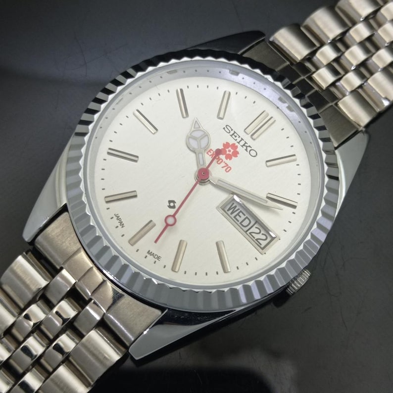 May include: A silver-tone Seiko wristwatch with a fluted bezel and a metallic link bracelet. The off-white dial features silver baton indices, a day-date window at the three o'clock position, and a red cherry blossom logo with the text EXPO'70.
