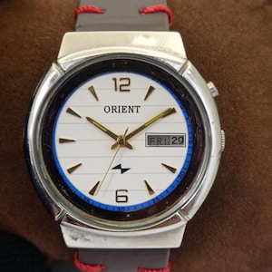 May include: Silver-toned Orient watch with a white face, gold hands, and a day-date display. The watch features a blue and black inner ring, and a gray leather strap with red stitching.