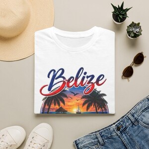 May include: A white t-shirt featuring the word Belize in a blue and red gradient script font. Below the text is a circular graphic depicting a tropical sunset scene with palm trees, a small hut, a sailboat on the water, and birds in flight.