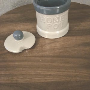 May include: A cream-colored ceramic honey pot with a gray rim and lid. The words "HONEY POT" are embossed on the front. The lid is off to the side, revealing the interior of the pot. The pot sits on a wooden table.