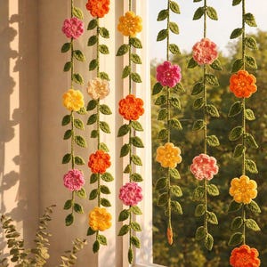 May include: Five vertical hanging garlands featuring crocheted green vines with small leaves and multi-colored flowers in shades of yellow, orange, pink, and cream. The floral strands are suspended in front of a window, creating a decorative display.