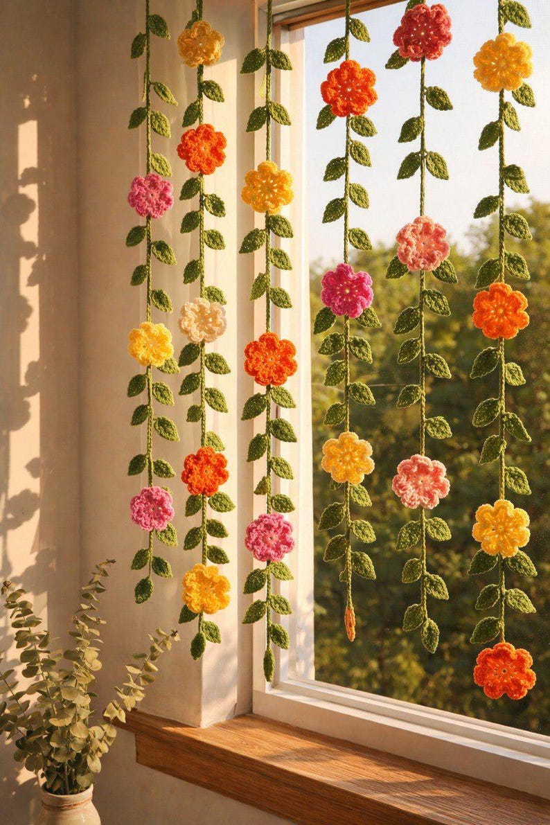 May include: Five vertical hanging garlands featuring crocheted green vines with small leaves and multi-colored flowers in shades of yellow, orange, pink, and cream. The floral strands are suspended in front of a window, creating a decorative display.