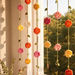May include: A set of vertical hanging garlands featuring crocheted flowers in shades of yellow, orange, pink, and white. Each strand is connected by a green yarn vine with small crocheted leaves, suspended in front of a window frame.
