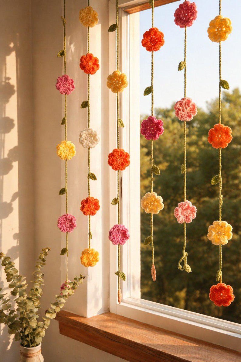 May include: A set of vertical hanging garlands featuring crocheted flowers in shades of yellow, orange, pink, and white. Each strand is connected by a green yarn vine with small crocheted leaves, suspended in front of a window frame.